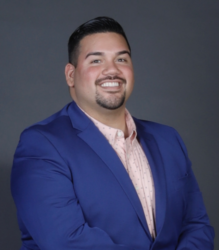 man in suit smiling, blue jacket, pink shirt dark hair with goatee